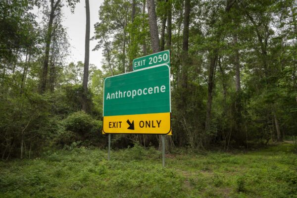 A freeway exit sign that reads "Exit 2050 Anthropocene. Exit Only" sits in a wooded clearing.