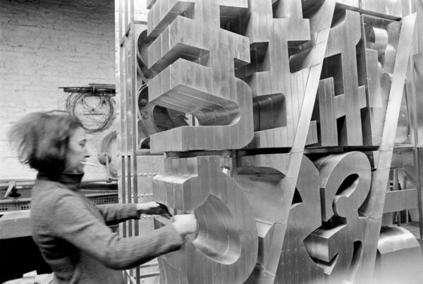 A black and white photograph of artist Chryssa adjusting a large sculpture.