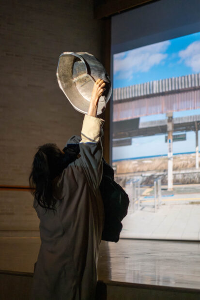 Image of an aartist performing in front of a video projection and holding a round bowl over her head