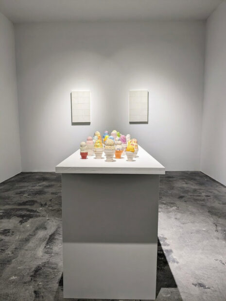 An installation image of an array of icecream sculptures sitting on a white table in a white walled gallery.