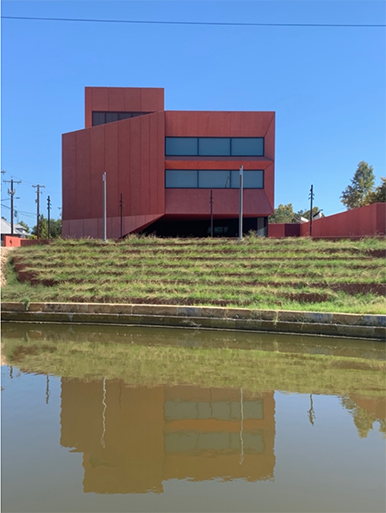 Un edificio rojo de dos pisos con ventanas largas y formas geométricas en la fachada, así como el pasto verde frente a él, se refleja en un cuerpo de agua en primer plano.