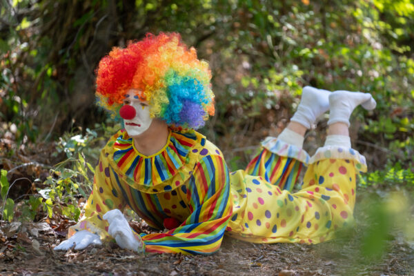 A sculpture of a realistic looking clown, with a rainbow wig, striped suit, and full makeup, lays on the ground.
