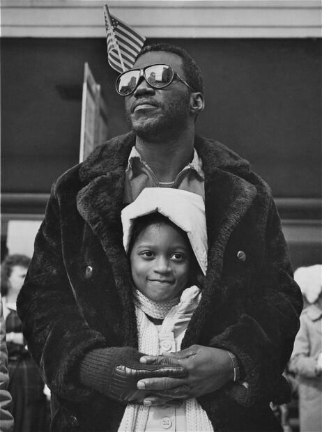 A black and white photograph of a father embracing his daughter while watching a parade.