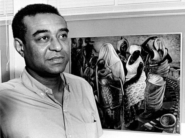 A black and white photograph of John Biggers with one of his paintings in the background.