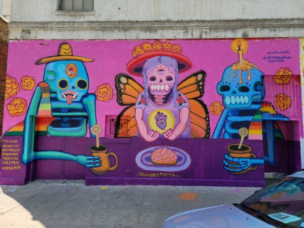 A brightly painted mural by Mauro de la Tierra featuring three skeleton figures surrounded by iconography related to Día de los Muertos.