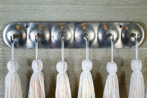 A photograph of a work of art by Jenelle Esparza featuring six white tassels hanging from metal hooks.