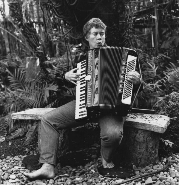Black and white image of a woman sitting on a bench outside, playing an accordion
