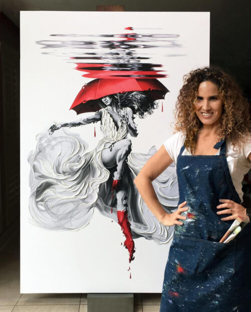 A photograph of artist Karina Llergo standing in front of a painting of a woman holding an umbrella and jumping in the air.
