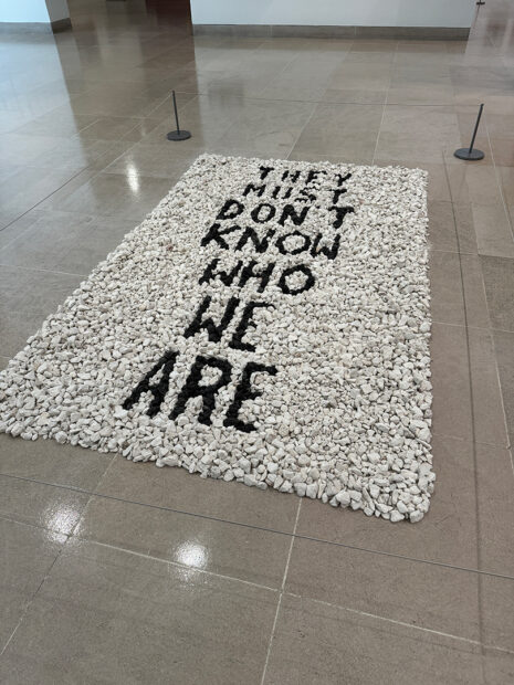 A photograph of an artwork made of black and white rocks with text that reads, "They Must Don't Know Who We Are." Artwork by Annette Lawrence.