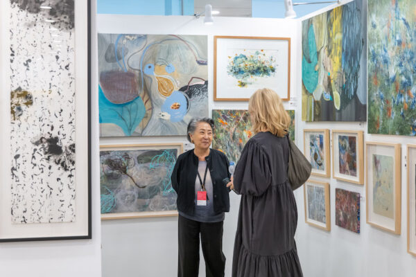 A photograph of two people standing in an art fair booth with paintings hanging on temporary walls.