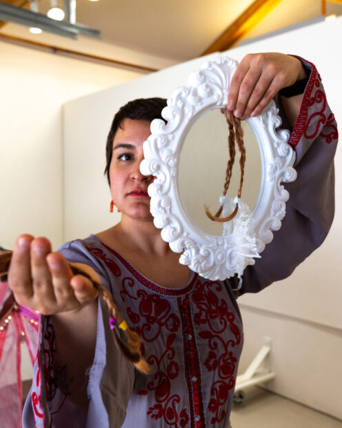 Photo of the artist holding a braid of hair with outstretched hand and a mirror