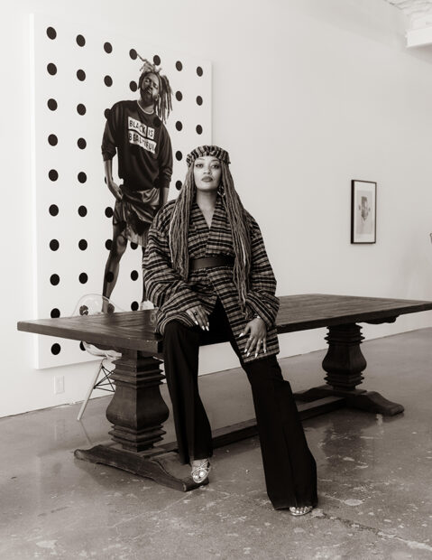 A black and white photograph of gallerist Daisha Board sitting on a table in front of a painting by Jeremy Biggers.