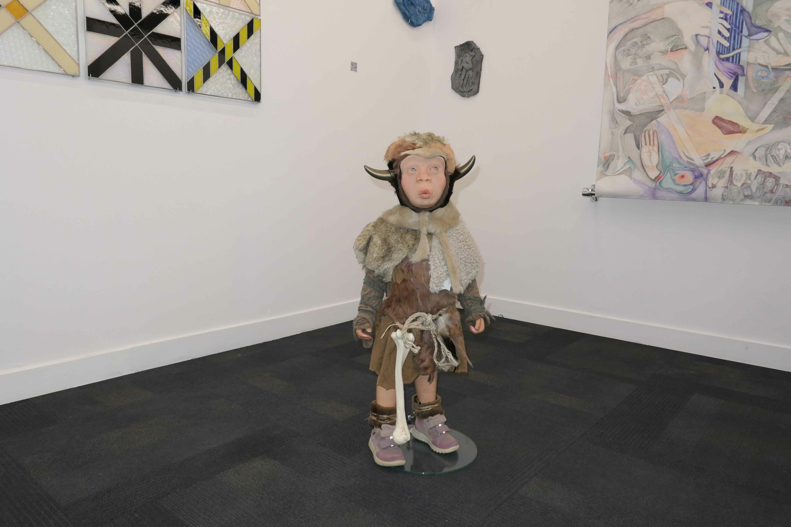 A sculpture of a small figure dressed in furs with a horned helmet stands before abstract paintings on the wall of a gallery.