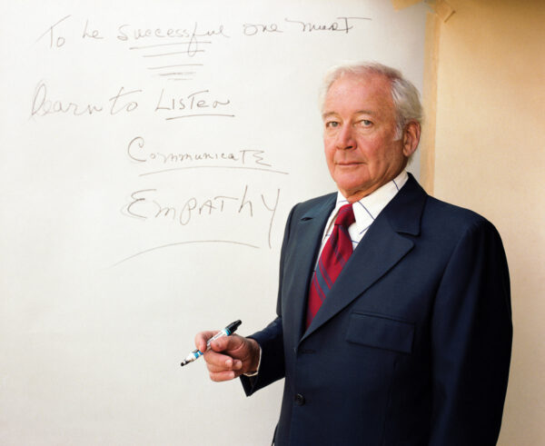 A man holding a marker and wearing a suit stands in front of a board that reads "to be successful one must learn to listen; communicate; empathy."