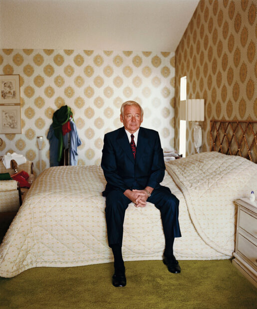 An older man wearing a suit sits on a bed. The room he is in has very patterned wallpaper.