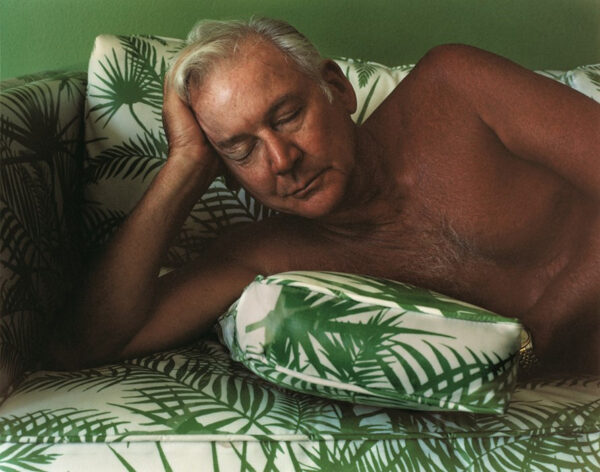A shirtless older man lays, with his eyes closed, on a leaf-patterned couch.