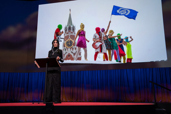 A photograph of artist and activist Nadya Tolokonnikova presenting a TED Talk with a photograph of her performance group Pussy Riot in the background.