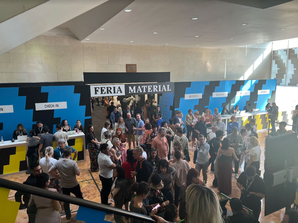 A group of people stand in the atrium of an art fair. Some people stand and talk in smaller groups, others pose for pictures, some people purchase tickets in line near the entrance doors.