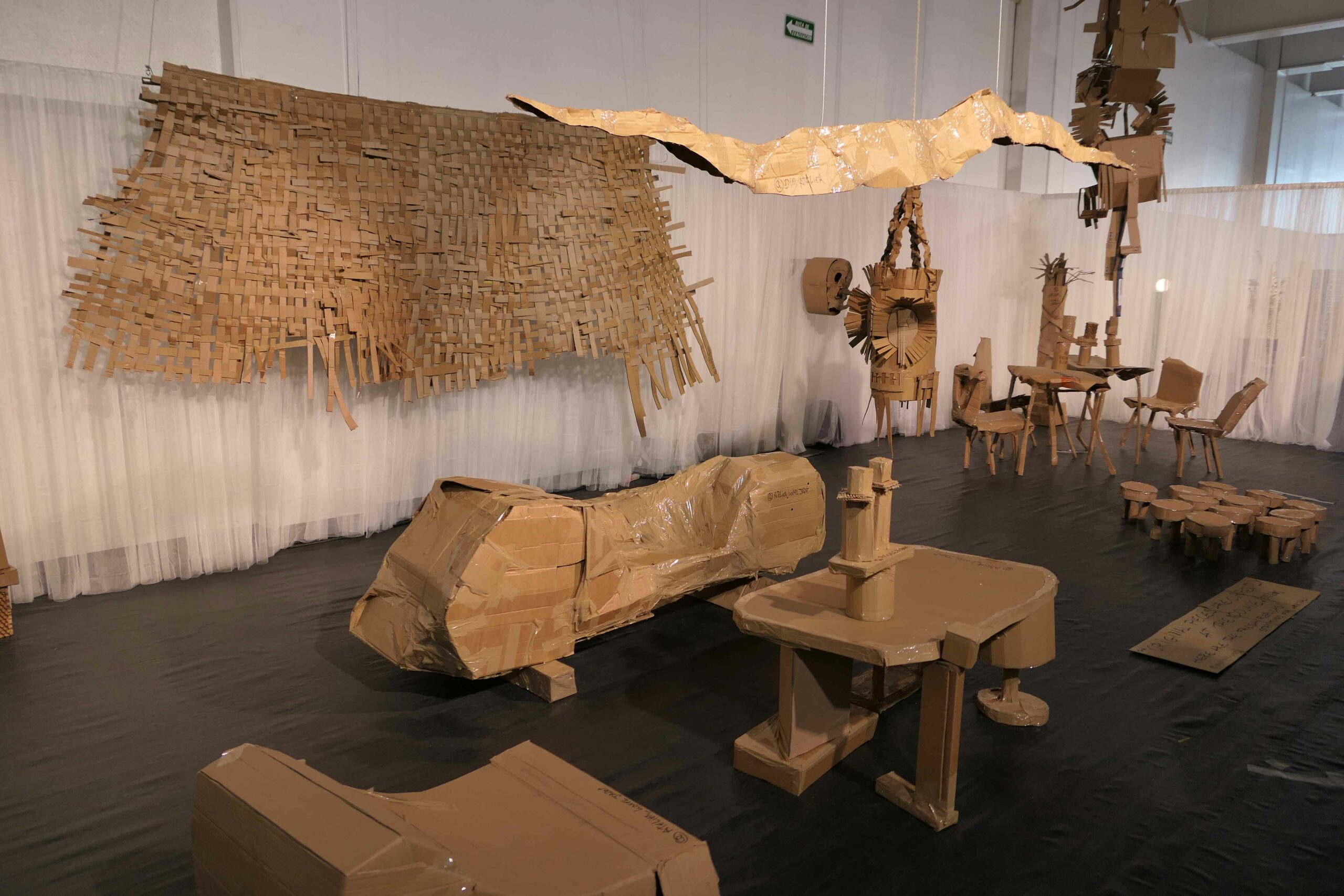 A room full of cardboard and tape sculptures. Some sculptures are abstract, some are furniture, such as chairs, tables and coffee tables. on the back wall is a large woven tapestry, also made from cardboard