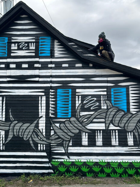 A photograph of muralist Kill Joy sitting on top of a house that she is painting.