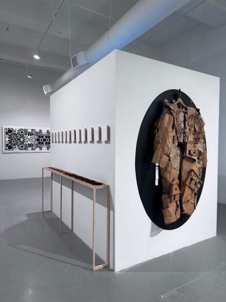 Installation view of square works on a wall on the right and a cardboard trench coat made on a black oval