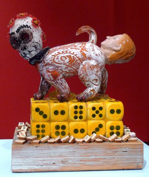 Glass sculpture of a cat body with a skull head and a doll head coming from the anus on a base of yellow and black dice