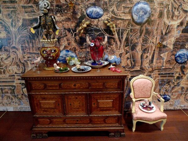 Installation with a wood bureau, an antique pink chair, both topped with small sculptures, and highly detailed wallpaper