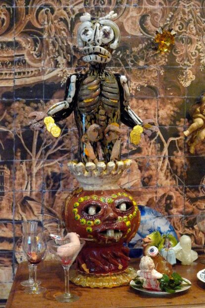 Detail of an installation of a skeleton on top of a ceramic sculpture with a face surrounded by smaller sculptures of fake dinner party pieces