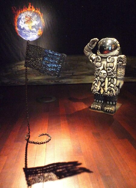 installation view of a sculpture of a cuatlique astronaut with a backdrop of planet earth on fire