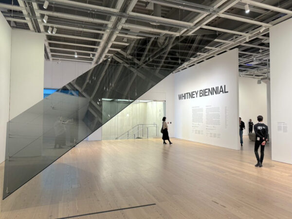 En una galería de paredes blancas con algunos visitantes flota inclinado un rectángulo negro y transparente. En una pared al fondo se puede leer “Bienal del Whitney” en inglés.