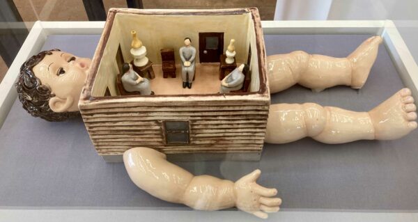 A ceramic sculpture of a baby whose torse is replaced by a small house with even smaller figures inside.