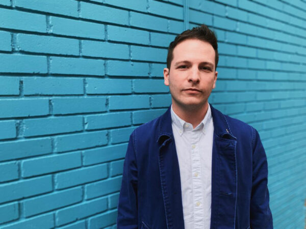 A photograph of curator Max Fields standing against a blue brick background.