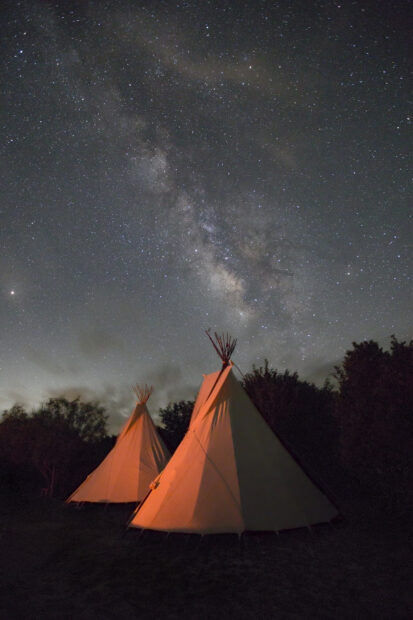 A photograph by Steve Goff featuring two teepees with an orange glow from a bonfire just out of frame against a night sky filled with stars and the Milky Way.