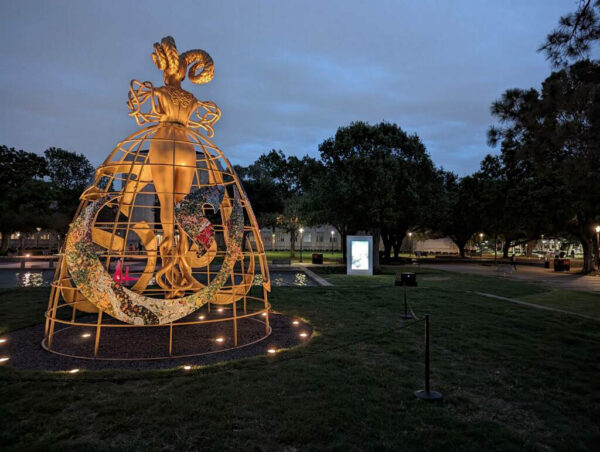 An evening photograph showing an outdoor installation of a large golden sculpture and a small screen in the background for a video work.