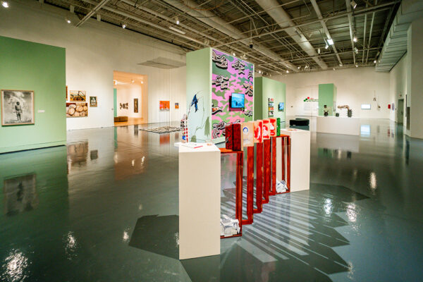 En una galería de techos altos y algunos muros blancos y otros color verde espárrago están dispersas distintas obras de arte de diversas técnicas y colores. 