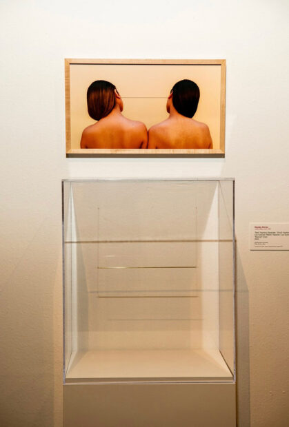 Sobre una pared blanca cuelga la fotografía de dos mujeres de espalda descubierta y cabello echado a un lado; están unidas por una línea que sale de los lóbulos de sus orejas. Debajo de esta fotografía se encuentra una vitrina que contiene una delgada barra de metal.