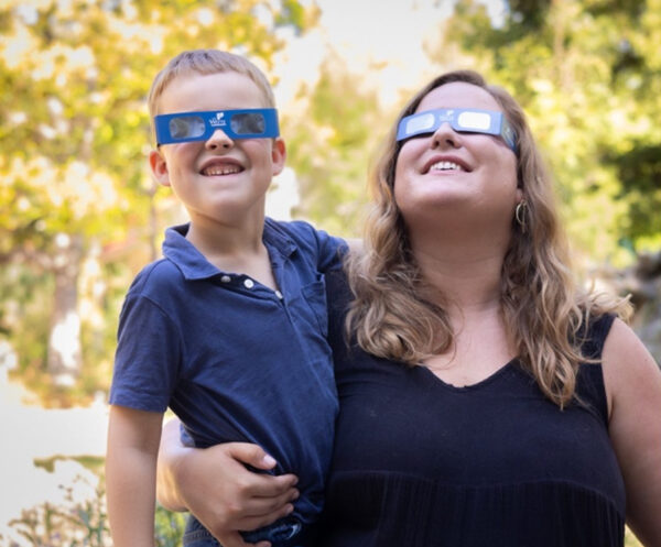 A photograph of a woman holding a young boy, both are wearing solar eclipse viewing glasses.