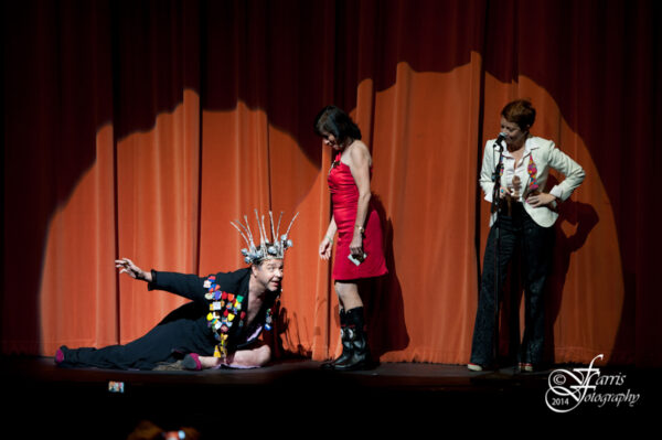 Photo of three actors on a stage, one kneeling on the ground wearing a silver crown