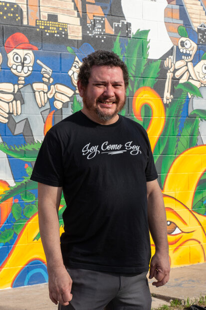A photograph of artist Juan Ramos standing in front of a mural he painted in San Antonio.