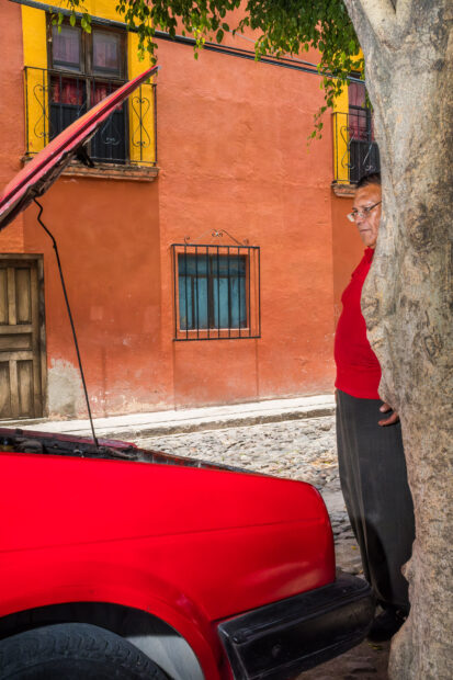 Photo of a man wearing a red tshirt standing behind a tree looking at a red car