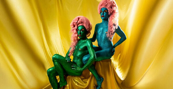 Two people with blue and green body paint wearing pink wigs pose before a golden backdrop.