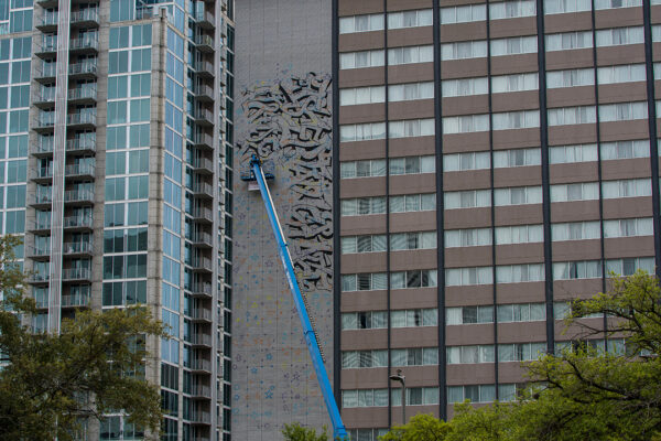 A photograph of Gonzo247 on a lift working on a large mural in downtown Houston.