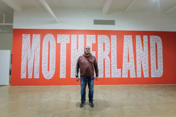 Photo of the artist standing in front of a mural of the words "motherland"