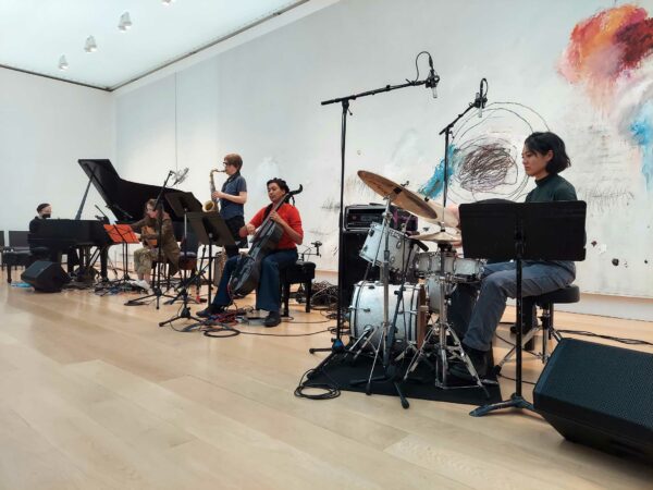 A quintet of musicians playing piano, guitar, saxophone, cello, and drums perform before a 30 foot Cy Twombly artwork.
