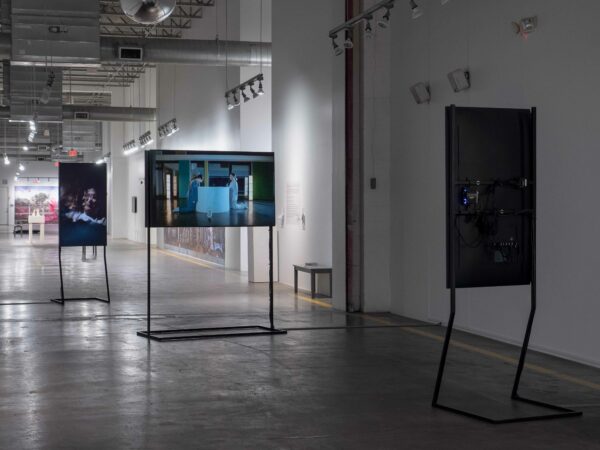 Installation view of three screens with images, and a large photo on a back wall in a hallway gallery