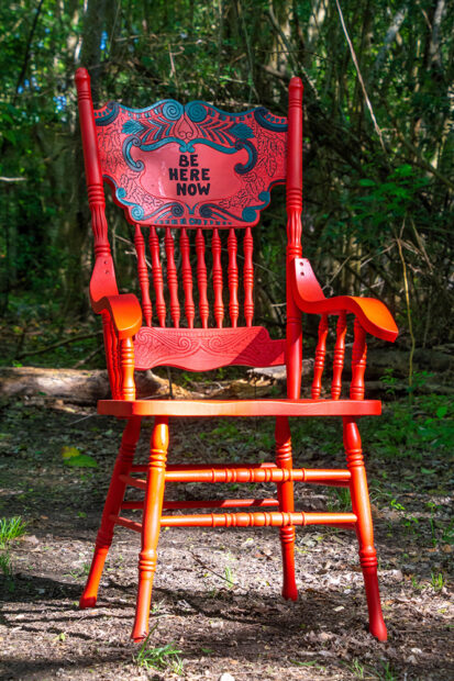 A photograph of a red chair with the words "Be Here Now" on its back.