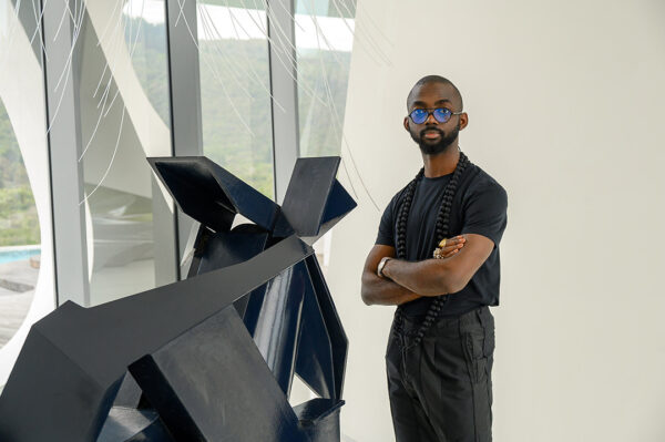 A photograph of artist Joseph Awuah-Darko standing next to a large abstract sculpture.