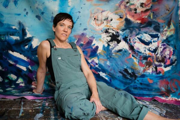 A person in coveralls sits before a large-scale abstract painting staring directly in to the camera.