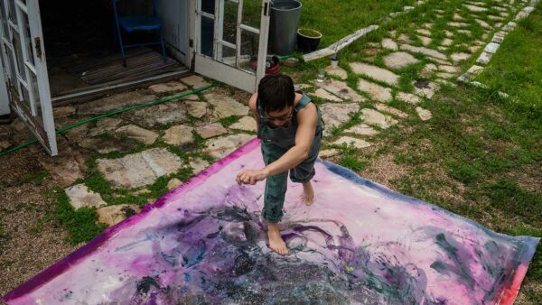 An unstretched canvas is spread out on the ground outside while the artist stands atop it.