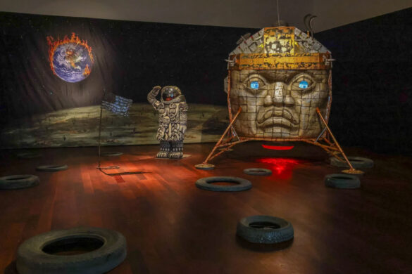 Installation view of a large Olmec head sculpture, an astronaut figure, and a moon like backdrop